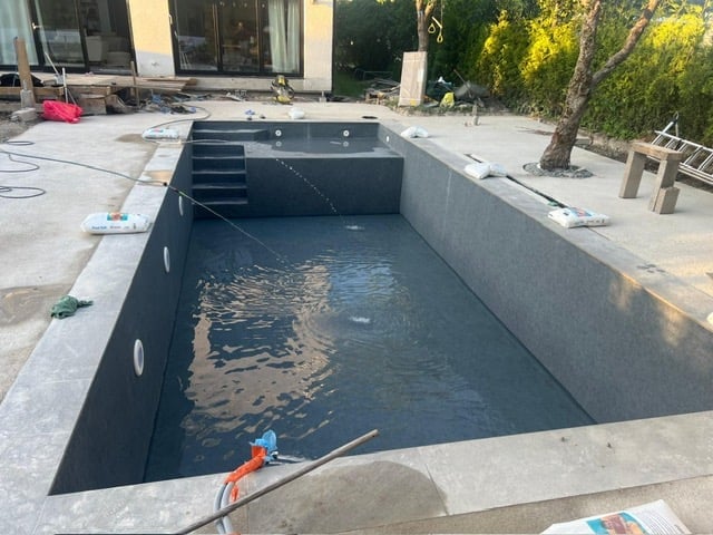 In-ground swimming pool under construction with concrete walls, filled with water, adjacent to a modern house and patio area